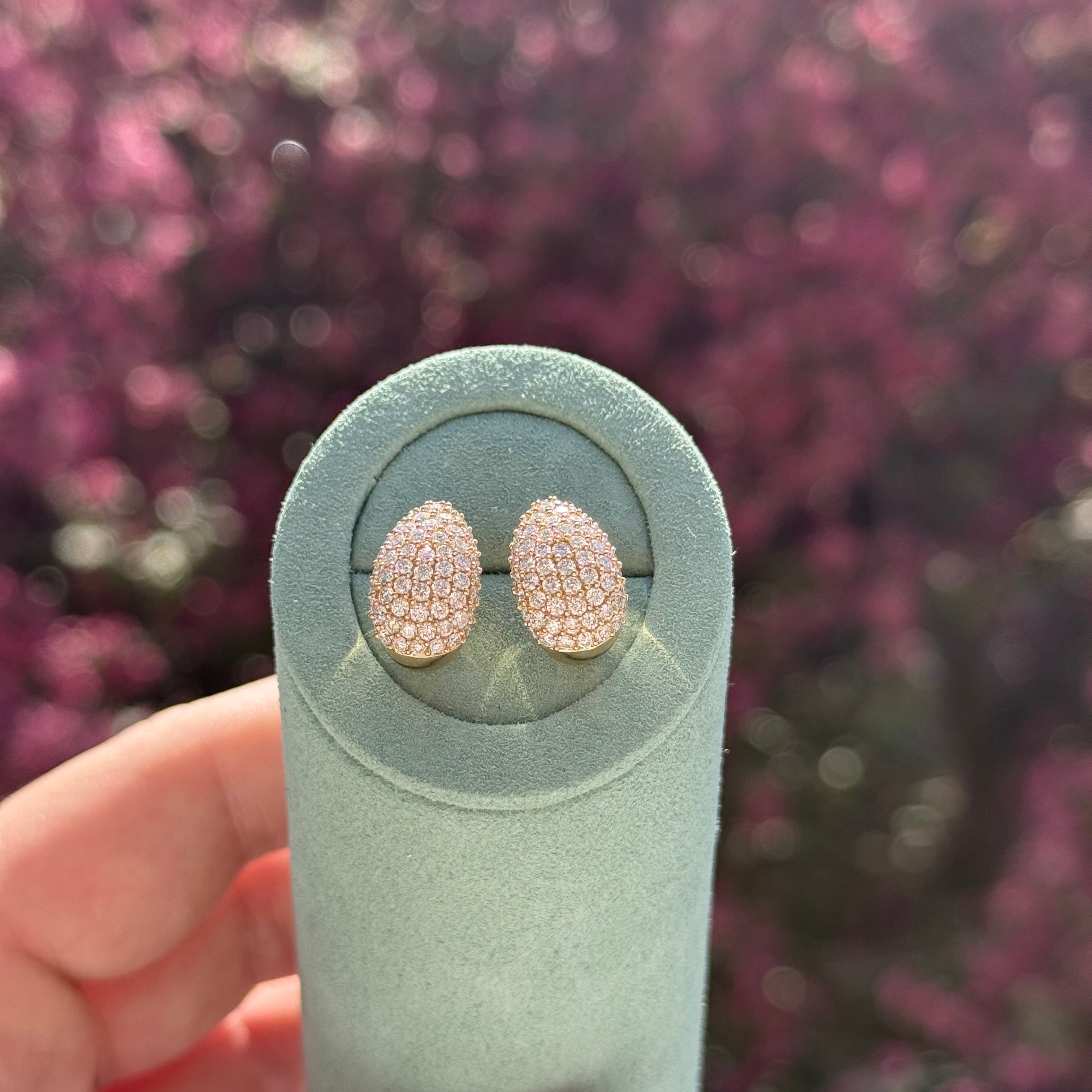 A pair of Diamond Teardrop Earrings with pavé-set diamonds in a 14k yellow gold setting, displayed on a round sage green jewelry stand held by a hand against a blurred background of pink blossoms.