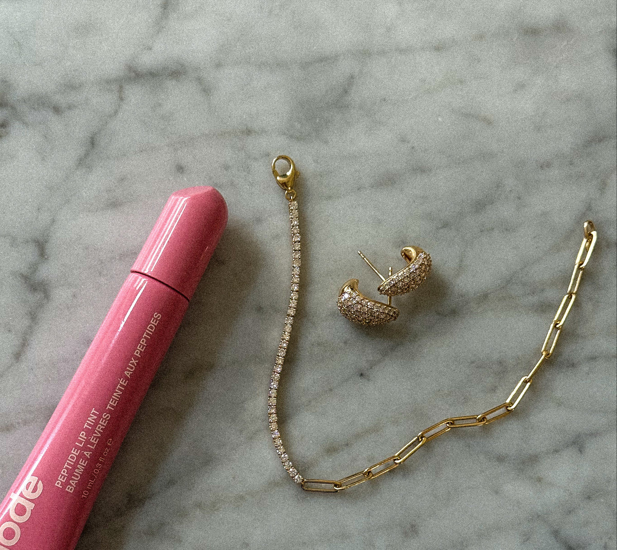 A pink tube of Rhode lip balm and a gold chain bracelet with a Diamond Teardrop Earrings, arranged on a white and gray marble surface.