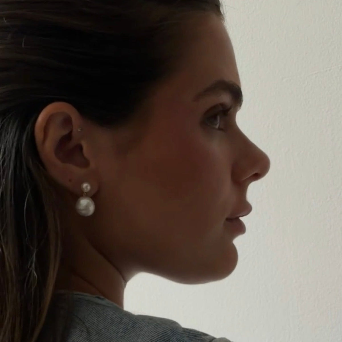 A close-up side profile of a woman wearing a pair of double-drop pearl stud earrings, shown against a plain off-white background.