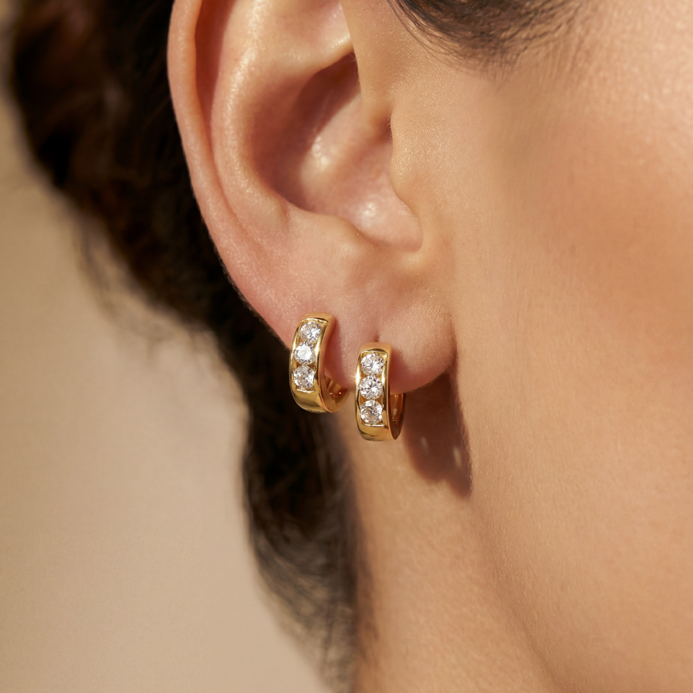 Close-up of an ear wearing gold hoop earrings with clear gemstones on a neutral background