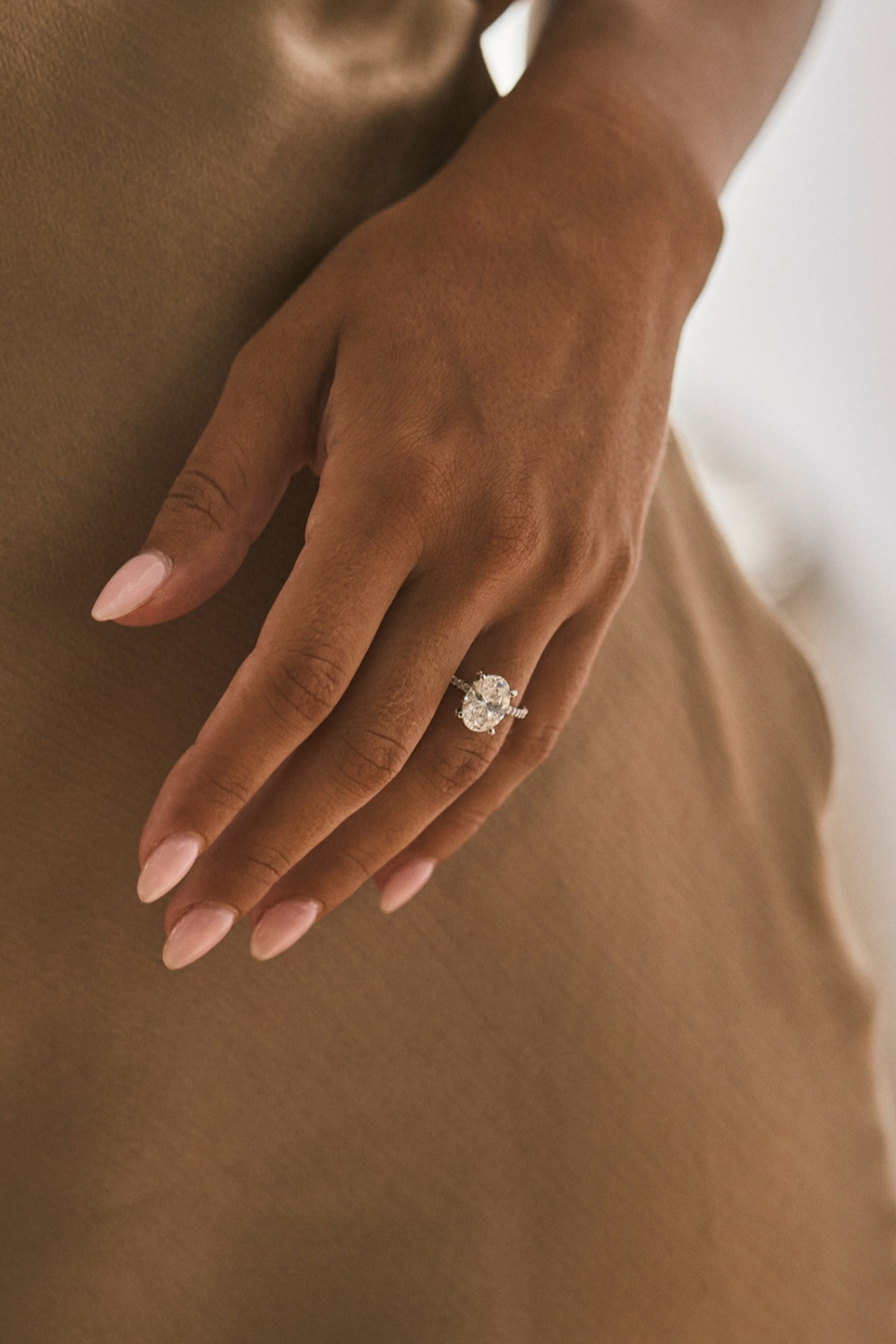 A close-up of a person's hand wearing a gold engagement ring with a diamond, set against a warm brown satin dress.