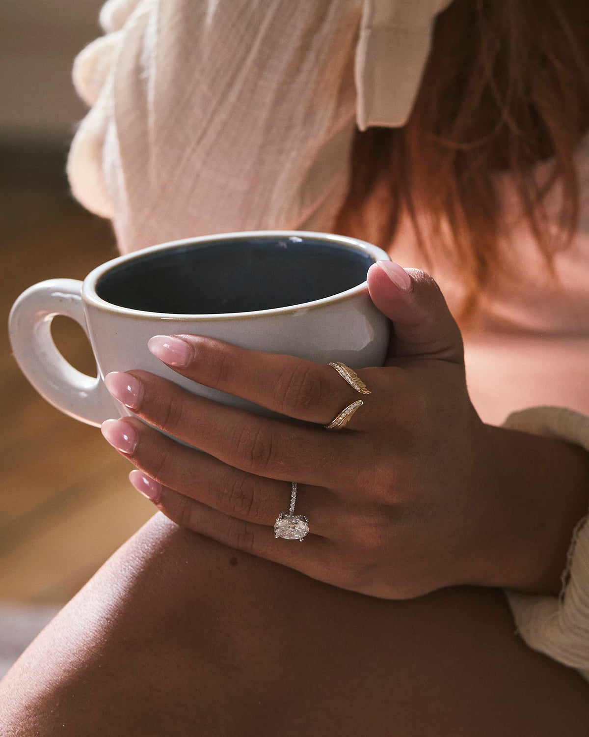 A close-up lifestyle shot of a person holding a white ceramic mug with a dark interior. The person is wearing a silver ring with a single round-cut clear gemstone on the ring finger, along with two gold bands on the middle and ring fingers.