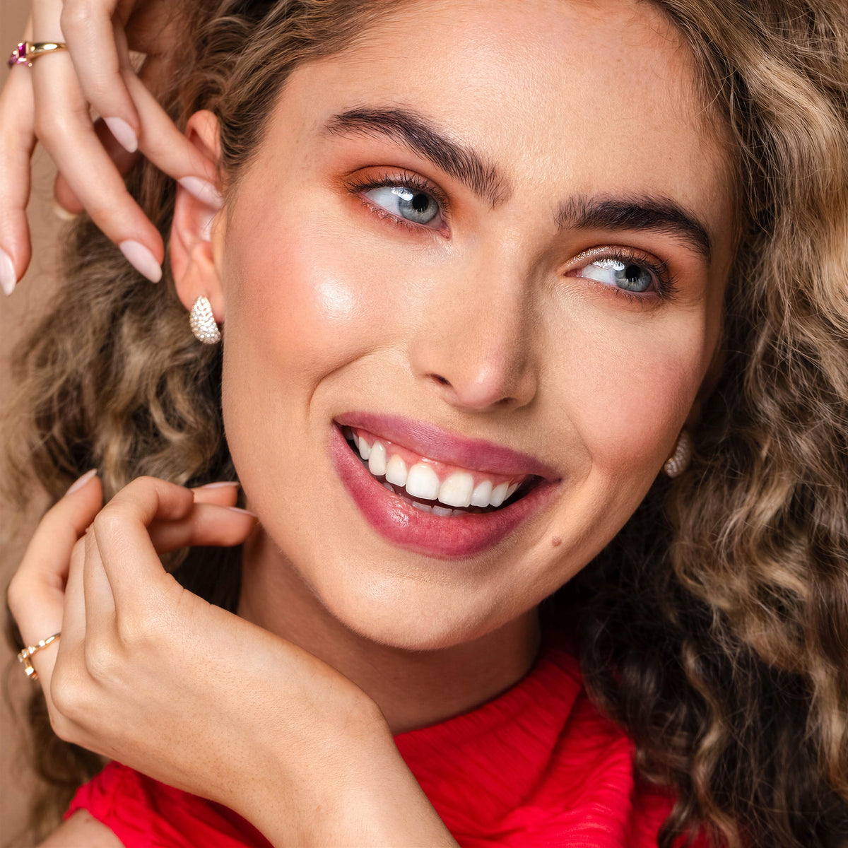 A close-up portrait of a smiling woman with curly brown hair wearing Diamond Teardrop Earrings and a gold ring, against a warm neutral background.