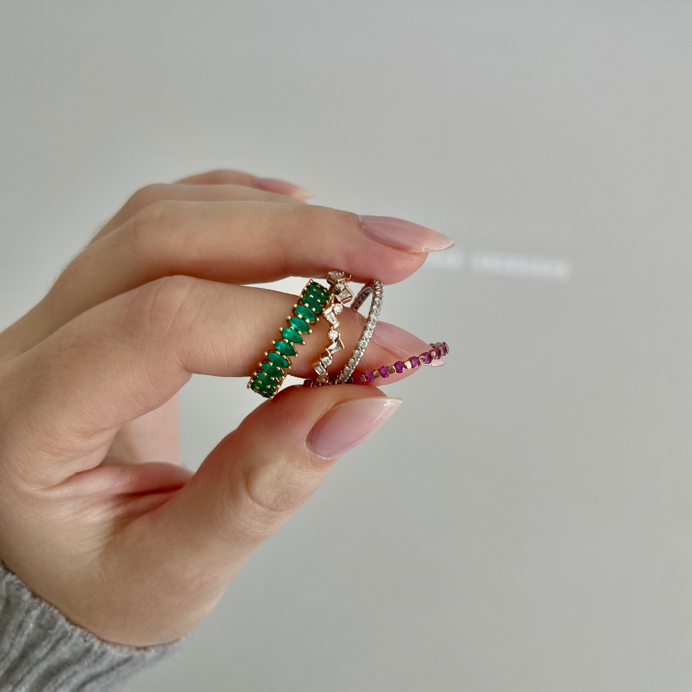 Hand holding two rings with green and red gemstones against a neutral background