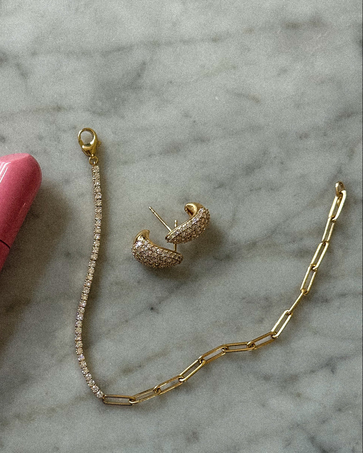 A pink tube of Rhode lip balm and a gold chain bracelet with a Diamond Teardrop Earrings, arranged on a white and gray marble surface.