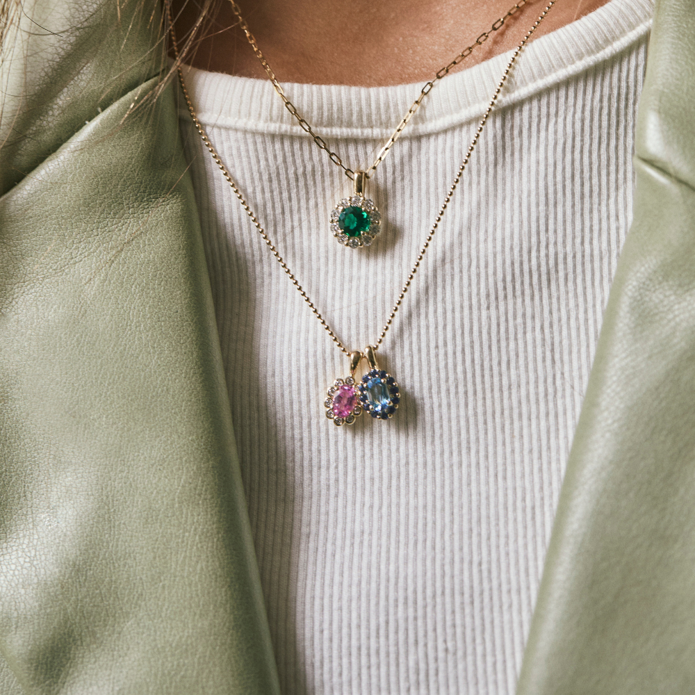 A close-up of a person wearing layered gold necklaces over a white ribbed top and sage green jacket. The necklaces include a single round green gemstone pendant and a double-drop cluster pendant with pink, blue, and green gemstones.