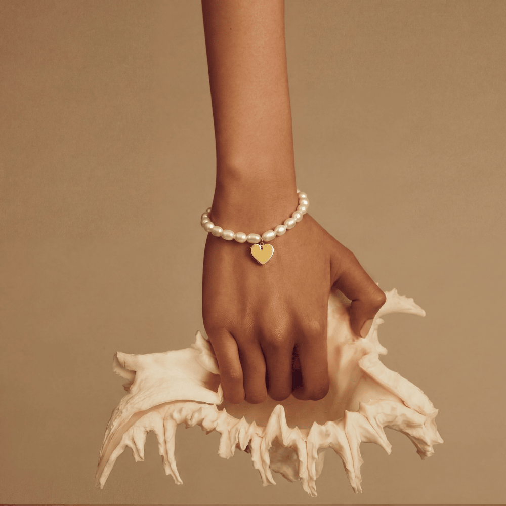 Hand wearing a pearl bracelet and gold heart charm on a beige background