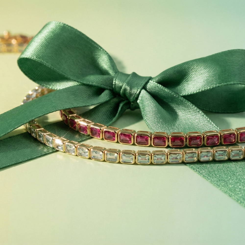 A close-up of a gold-toned tennis bracelet with rectangular pink and blue topaz gemstones, tied with a large satin green ribbon bow on a pale mint background.