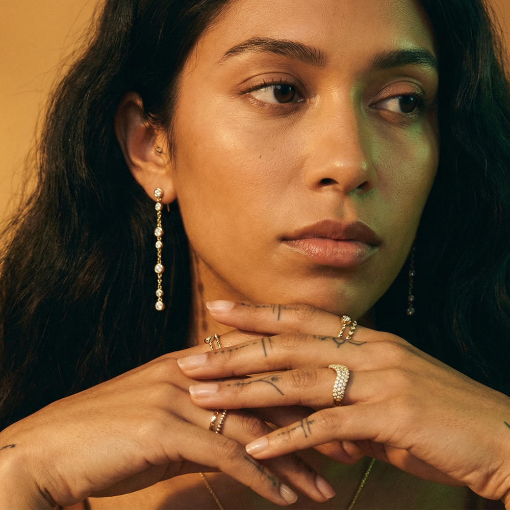 Close-up of a woman with jewelry, including diamond chain earrings and rings, against a warm-toned background.