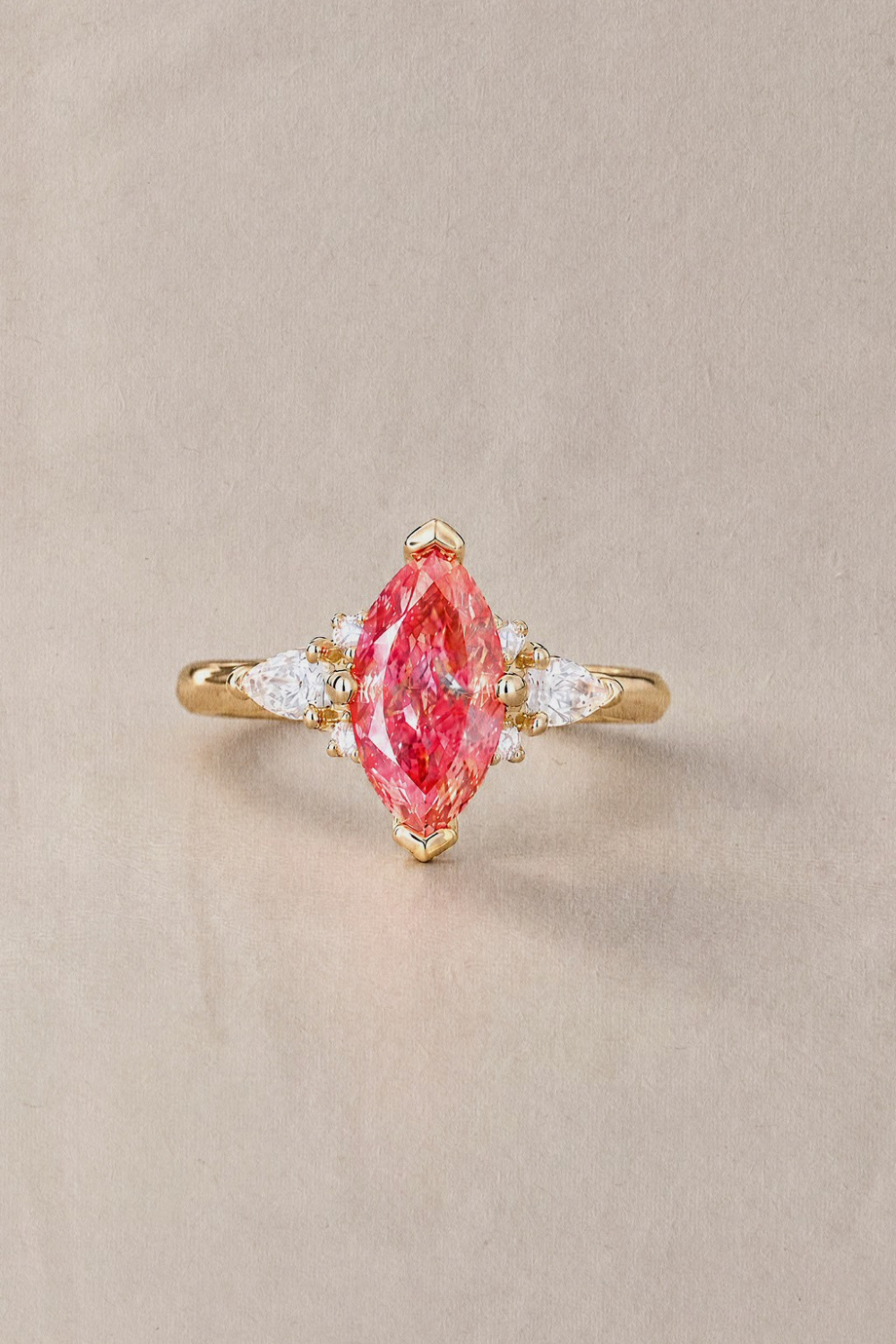 A gold ring featuring a marquise-cut pink diamond in the center, flanked by small diamonds on a warm beige background.